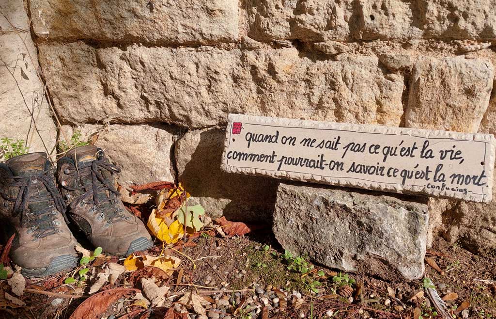Chaussures de marche pèlerin déposées devant une pierre gravée d'une citation de Confucius à Lauzerte sur le chemin de Saint-Jacques-de-Compostelle.